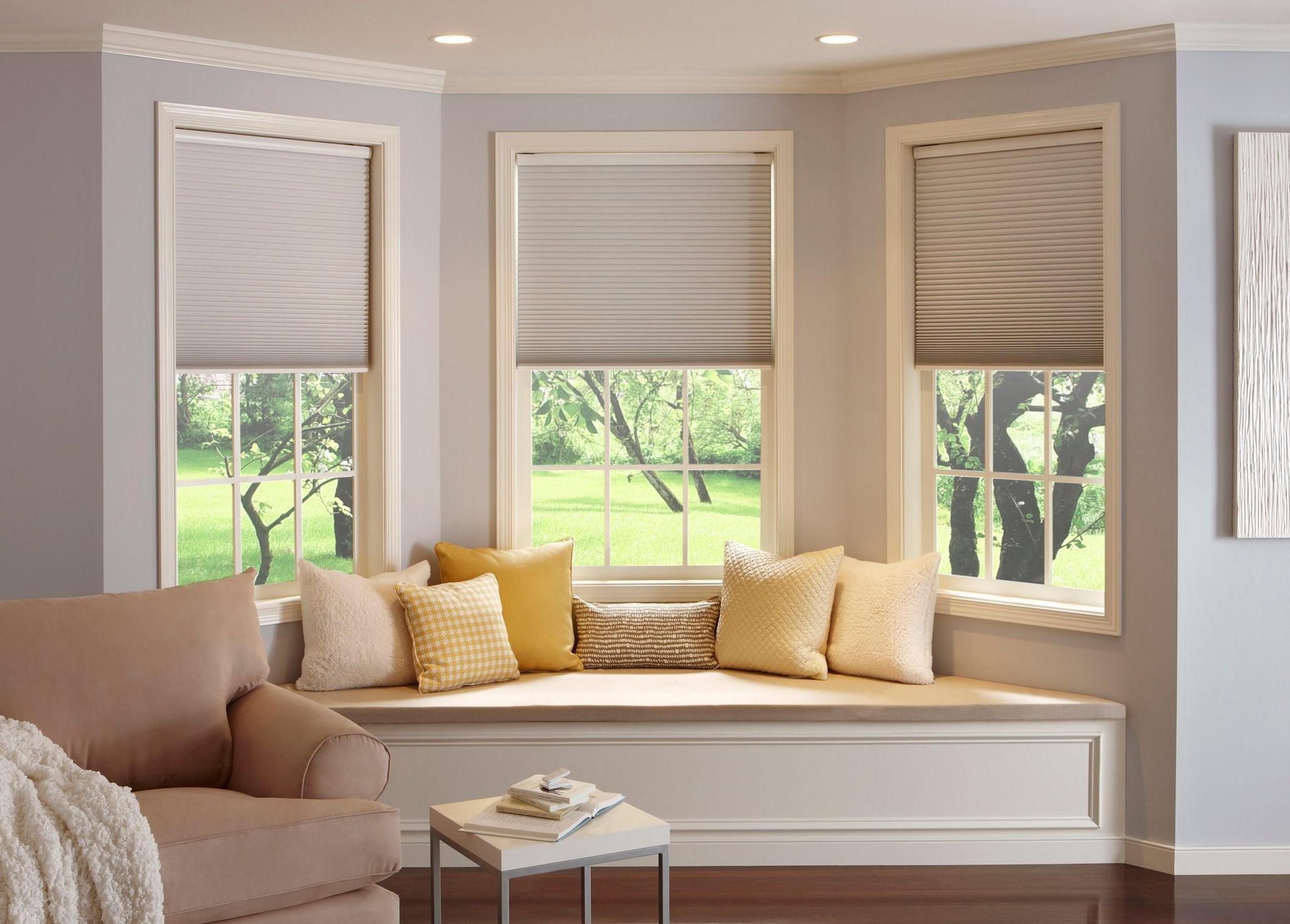 A cozy living room features a bay window seat with beige and yellow cushions. Partially drawn white shades let sunlight illuminate the space. A beige couch sits to the left, and a small white table is placed in front of the window seat.
